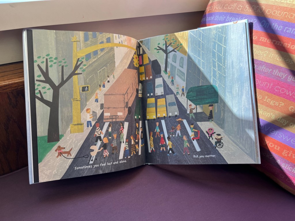 An image of the book You Matter opened up to a painting of a city with various people using a cross walk. The text reads, "Sometimes you feel lost and alone. But you matter."