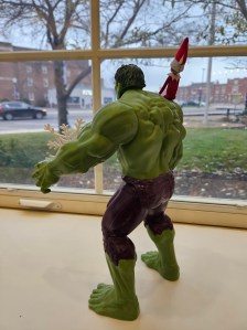 an incredible Hulk toy with a tiny Elf on his shoulder looks out the library window, appearing lonely
