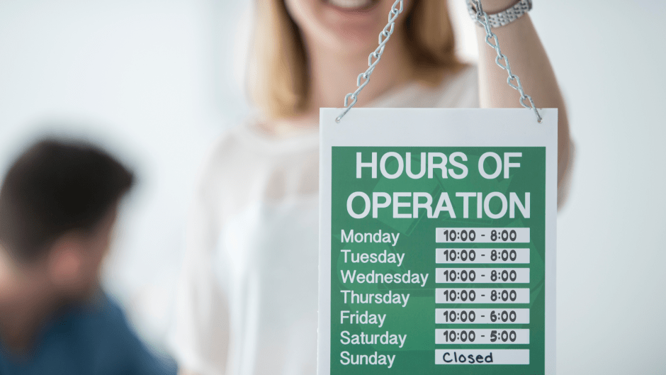 person holding a business hours sign showing the days of week and hours open each day.