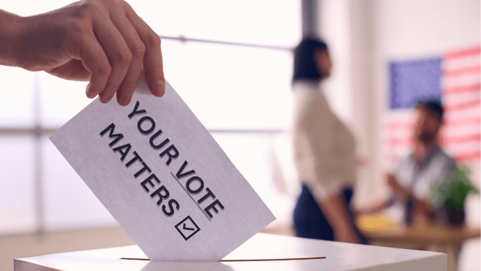 in the foreground, a hand holding a card labeled "your vote matters" inserts it into a ballot box. In the blurred background are an American flag and two other people.