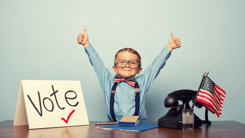 A smiling young boy in glasses, a blue shirt, suspenders, and a stars-and-stripes bow tie gives two thumbs up at a desk, next to a handwritten "Vote" sign with a red checkmark and a small American flag.